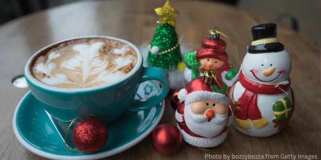 Coffee in a blue cup with saucer and several Christmas ornaments on the table