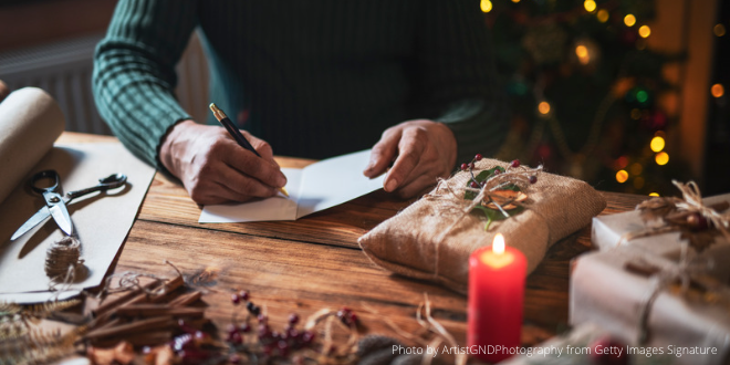 A man sitting at his table writing Christmas greetings in a card surrounded by Christmassy things