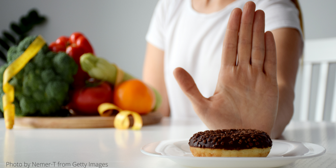 A woman siting in front of a plate of vegetables, holding her hand up to refuse a chocolate donut.