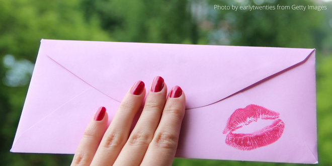 A female hand with red nail varnish holding a pink envelope with a red lipstick print on the back