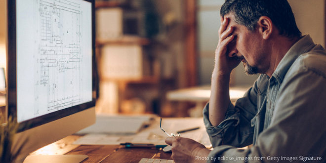 A man sitting in front of his computer in the evening looking overworked (with his head in his hand)