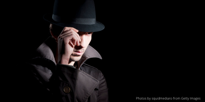 A man dressed in a black trenchcoat, wearing a black hat in front of a black background