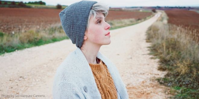 A young woman wearing a blue hat, standing on a stony path and looking over the fields as if she's day dreaming