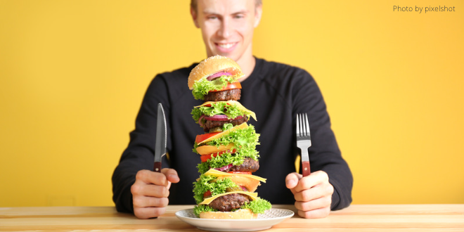 A young man sitting in front of a huge hamburger, with a knife and fork in each hand