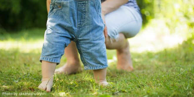 The legs of a baby taking his/her first steps on the grass, supported by the mother in the background