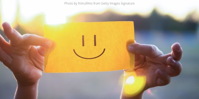 Hands holding up a yellow piece of paper against the sun, with a smiley on it