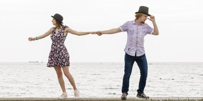 A woman and a man standing next to each other with their arms out stretched, holding each others hand, with the sea behind them