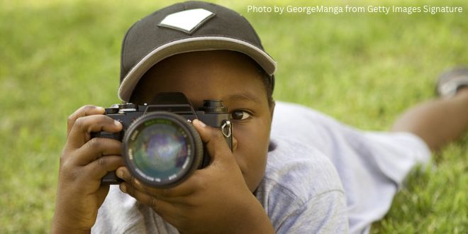 A young boy lying in the grass with a camera in front of his face