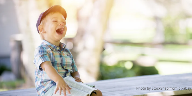 A young boy sitting on a bench outside, with a book on his lap, laughing