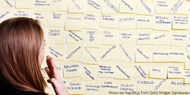 A woman standing in front of a board covering in Post-Its with business buzzwords and jargon