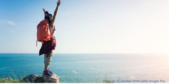 A woman wearing a rucksack on her back and standing on rock facing the sea, with her arms in the air