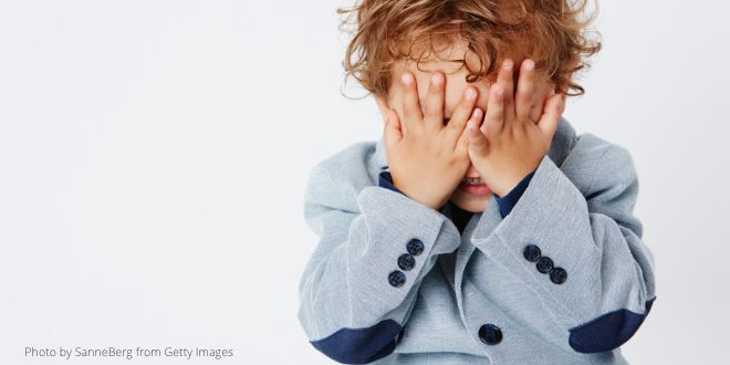 A young boy wearing a light blue jacket and hiding his face in his hands