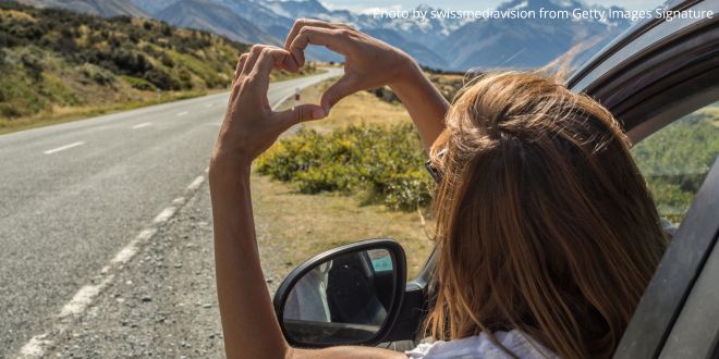 A young woman looking out of the window of a car and making a heart shape with her fingers