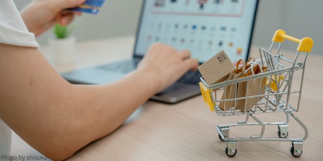 A close-up of a woman's arm in front of laptop, a credit card in her other hand, and a small shopping trolley filled with shopping bags in the foreground