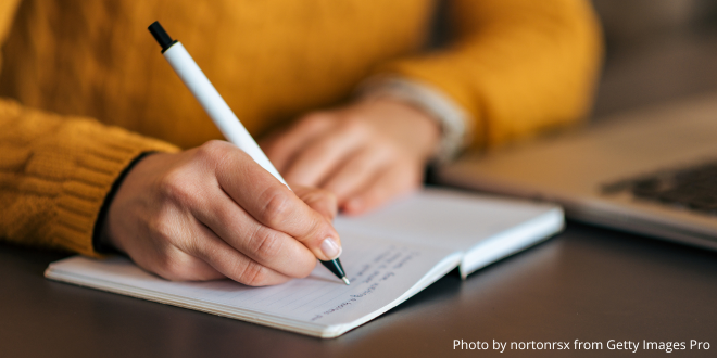 Close-up of a person in a yellow jumper writing in a notebook with pen in the right hand