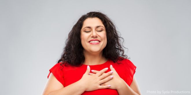 A woman with long dark hair and wearing a red dress, holding her hands over her heart